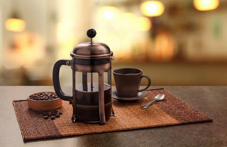 Bronze French press filled with coffee on a cozy cafe table with cup, spoon, and coffee beans