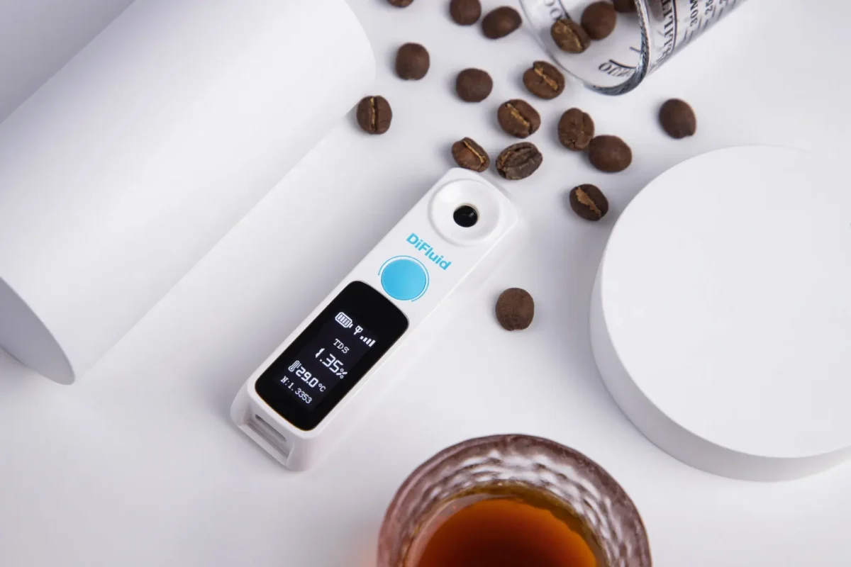 رفرکتومتر قهوه DiFluid در حال نمایش عدد TDS روی میز سفید همراه با دانه‌های قهوه