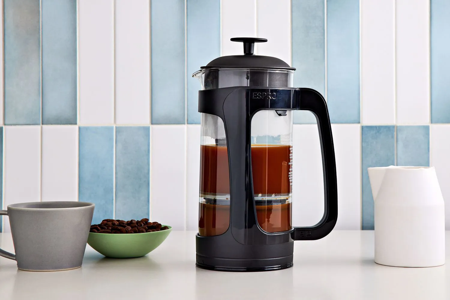 Modern black French press with fresh brewed coffee beside a gray mug and bowl of coffee beans