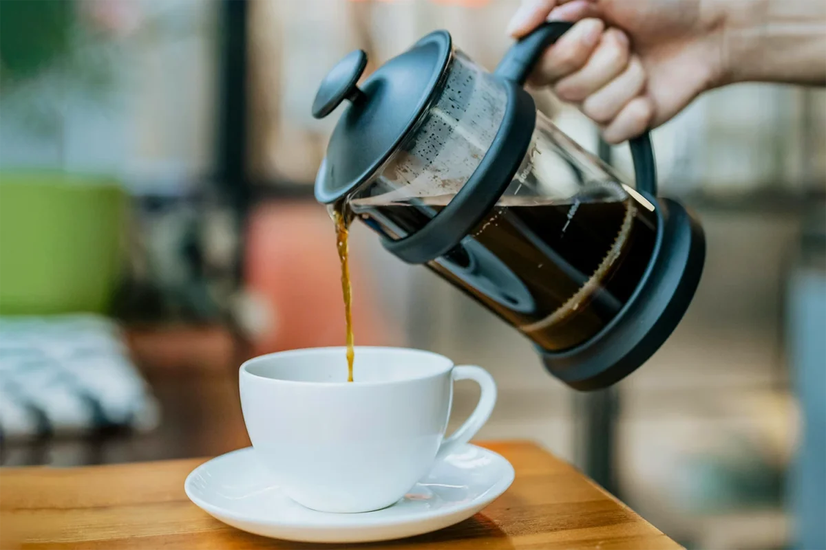 Fresh coffee being poured from a French press into a white cup on a wooden table